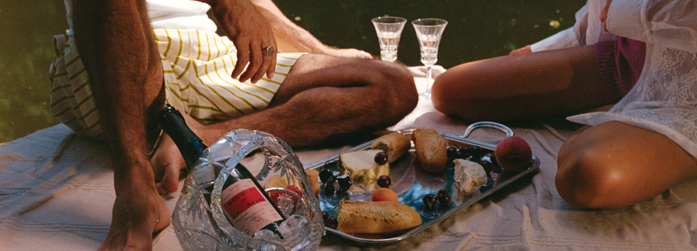 Two people sitting on a blanket with food and drinks outdoors