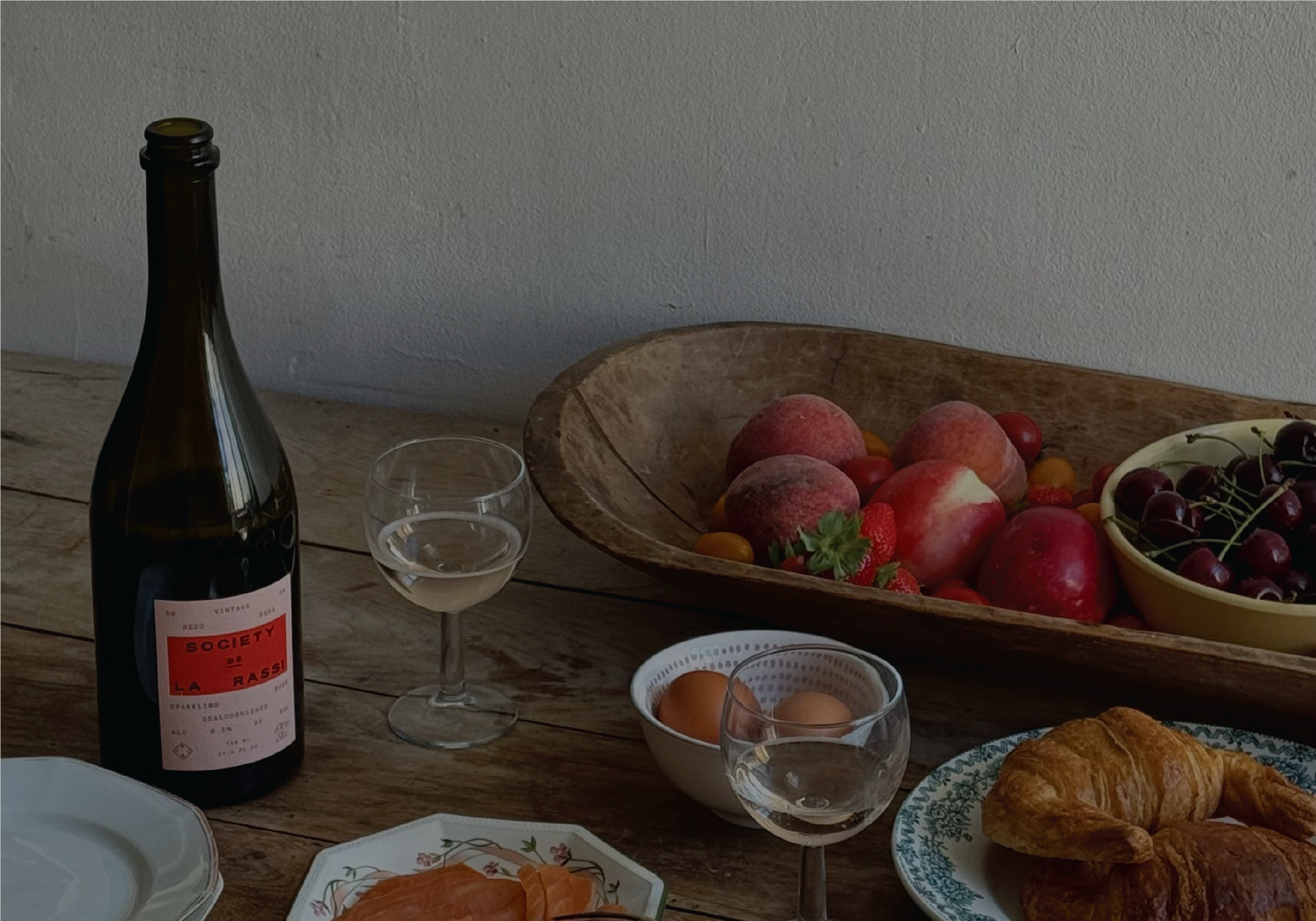 Wine bottle, glass, fruit bowl, eggs, and croissants on a wooden table.
