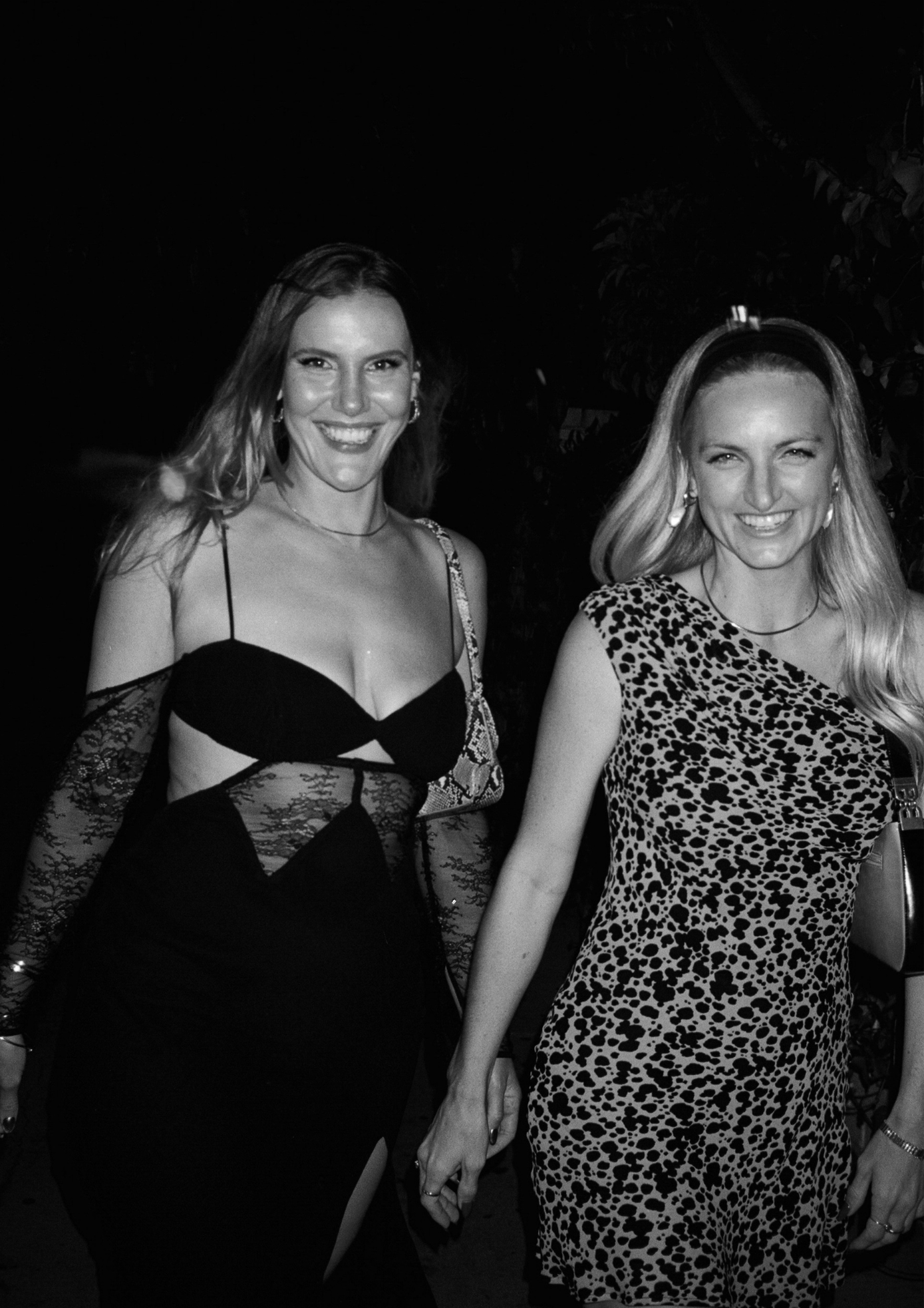 Two women standing together at an event, one in a black dress with cut-out design and the other in a leopard print dress.