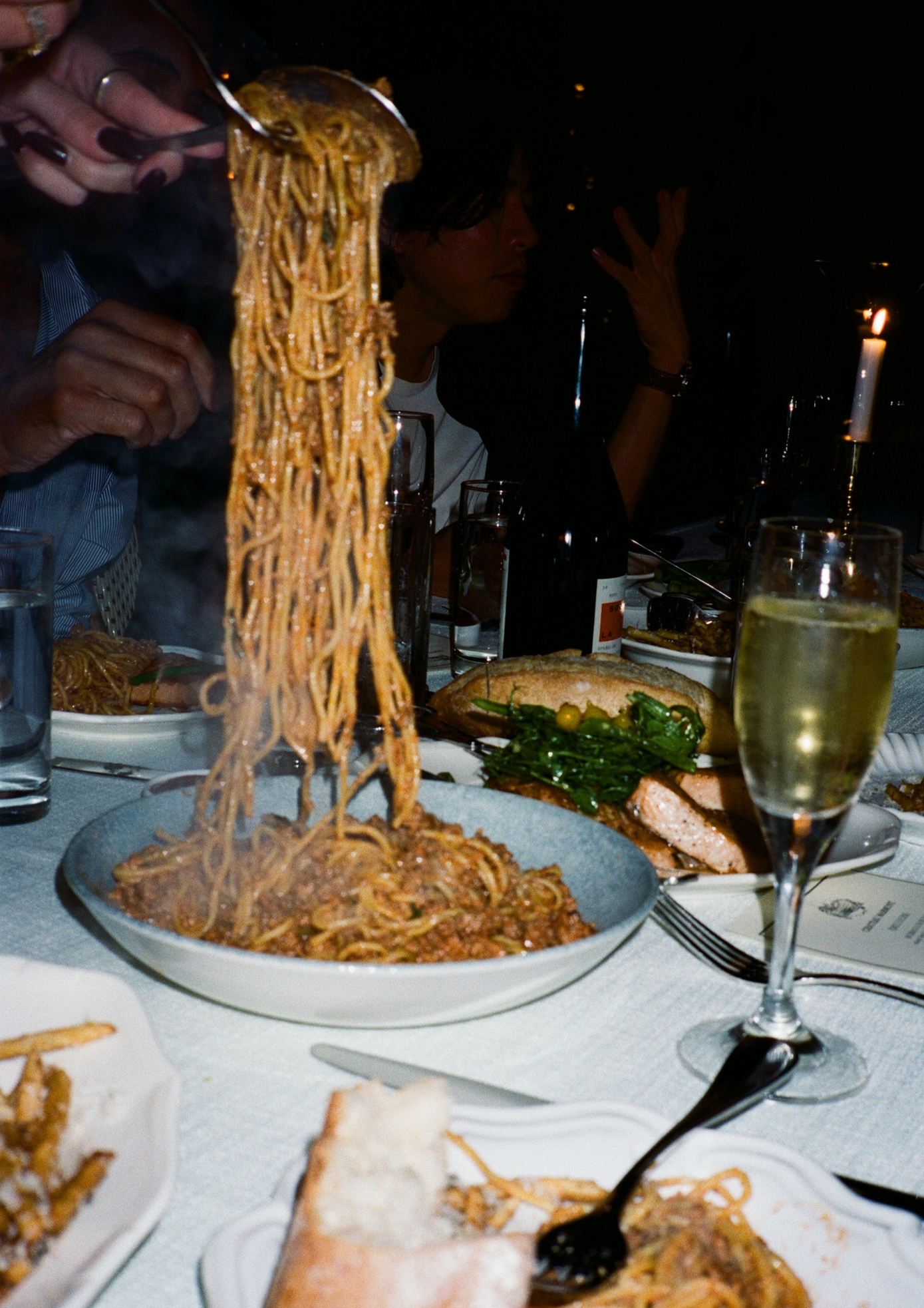 Spaghetti being lifted from a bowl with a fork, set on a dining table with wine and bread.