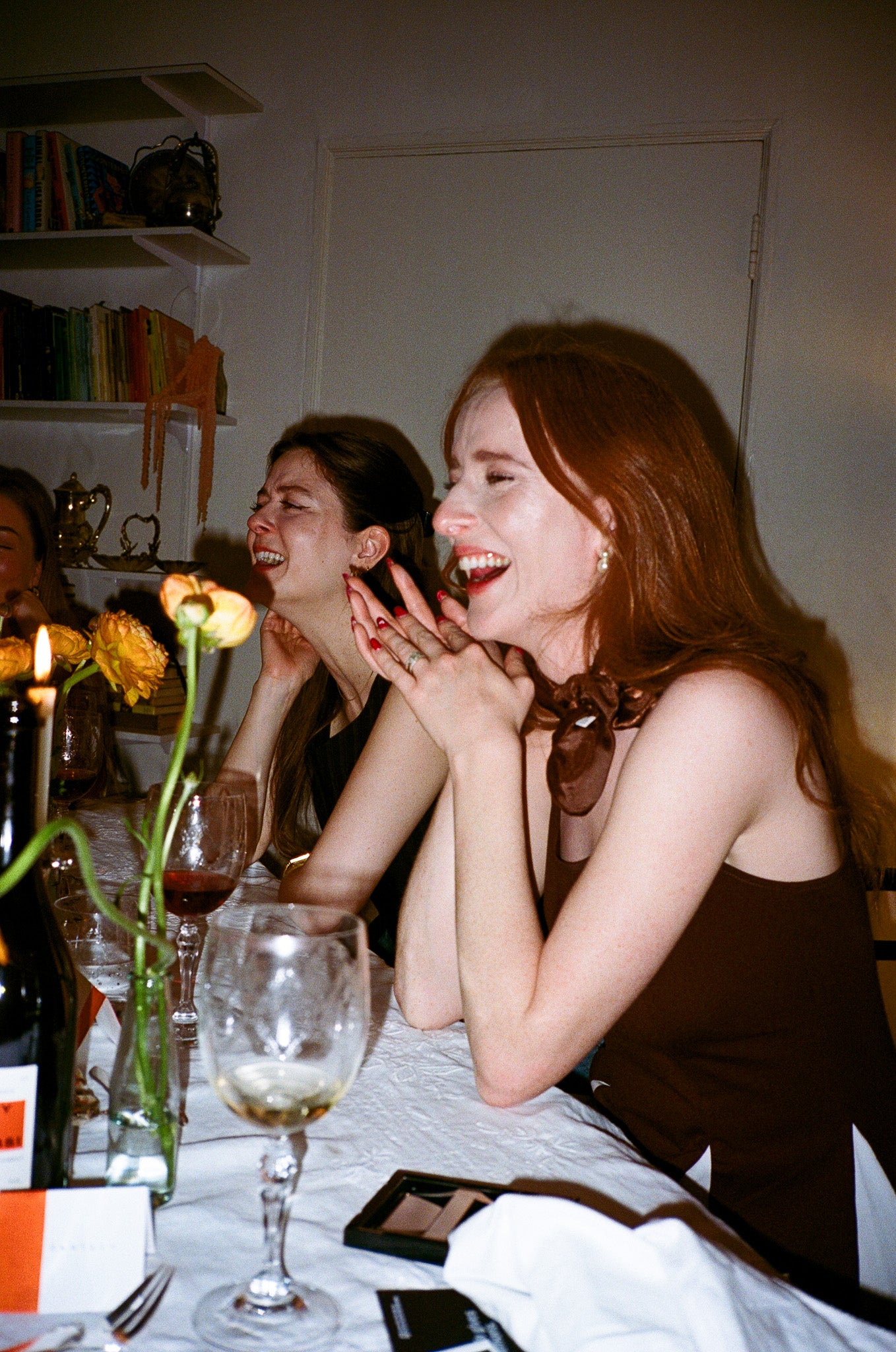 Two women sitting at a dinner table with wine glasses and a candle, laughing and smiling.